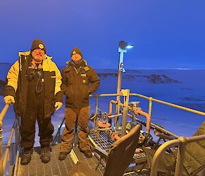 Two men stand on a wooden platform in low light
