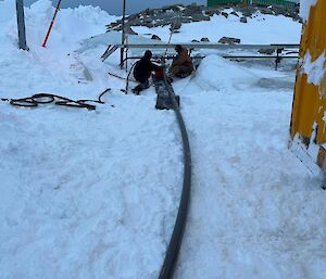 Two people sit in the snow, with a fuel hose stretching out behind them