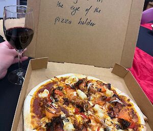 An open pizza box, with the phrase 'Beauty is in the eye of the pizza holder' written on the inside of the lid is open with a cooked pizza inside. A glass of red wine sits beside it