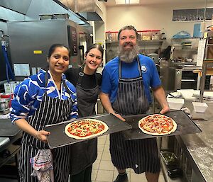 Three people wearing aprons stand in a kitchen holding tracys with pizzas ready to go in the oven