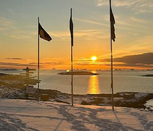 The flag poles with flags gently waving in the breeze stand in a snowy landscape with calm ocean visible behind and a sun just above the horizon