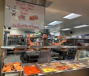 A man in a brightly coloured shirt and a blue and hite striped apron gives a double thumbs up, standing behind a bain marie full of food