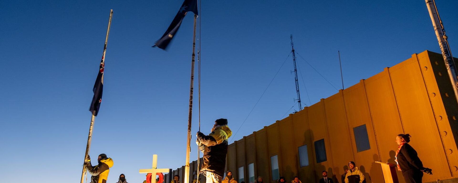 A group of people wearing thick jackets watch on respectfully as the Australian and New Zealand Flags are lowered, in front of a square yellow building with a pale dawn sky visible behind