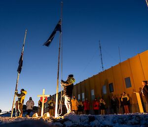 A group of people wearing thick jackets watch on respectfully as the Australian and New Zealand Flags are lowered, in front of a square yellow building with a pale dawn sky visible behind
