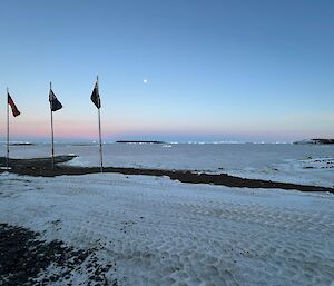 The flag poles with flags gently waving in the breeze stand in a snowy landscape with calm ocean visible behind and the moon visible in a pale blue sky