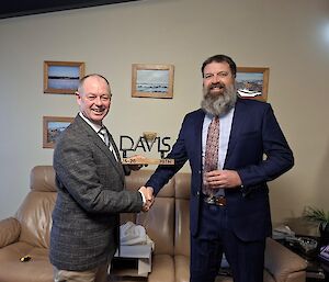 Two men wearing suit jackets are smiling at the camera whilst one is presented a gift of a wooden base supporting a metal sculpture of the word Davis