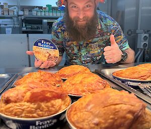 A man holding a Fray Bentos tin, stands smiling near five hot pies with a puff pastry top and Fray Bentos can on the bottom