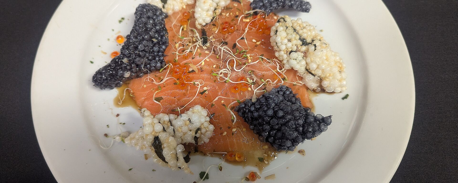 A white dinner plate holding thinly sliced salmon crudo dressed with yuzu ponzu, topped with sprouts and salmon roe, and surrounded by black and white crispy wakame puffs for contrast and texture.