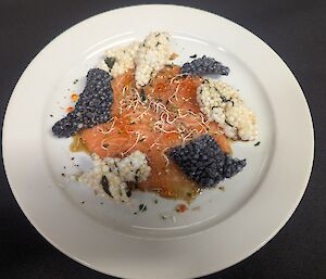 A white dinner plate holding thinly sliced salmon crudo dressed with yuzu ponzu, topped with sprouts and salmon roe, and surrounded by black and white crispy wakame puffs for contrast and texture.