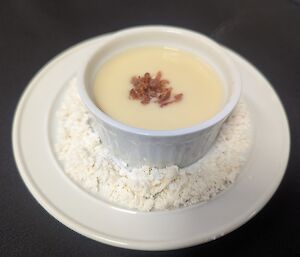 A small white ramekin filled with smooth, creamy parsnip velouté, garnished with a sprinkle of crispy bacon pieces and presented on a bed of fine white powder on a matching plate.