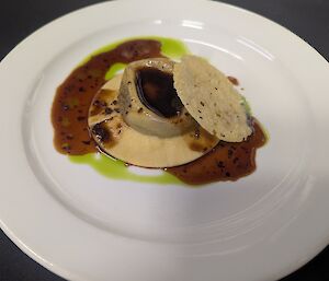 A large handmade oxtail raviolo served on a creamy sauce with rich truffle jus, bright green herb oil and topped with a crisp parmesan wafer on a white plate.