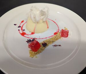 A smooth white cardamom pannacotta topped with a cloud of spun sugar, served with rosewater syrup, pistachio praline crumble and jewel-like pink jelly cubes on a white plate.