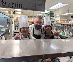 Three smiling chefs wearing white chef hats and uniforms pose together behind the kitchen pass in a commercial kitchen, with stainless steel equipment and workstations visible in the background.