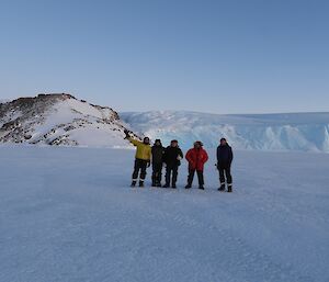 A photo of five expeditioners standing on the sea ice, with a rocky outcrop and ice walls in the background