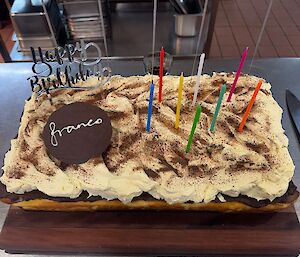 A very large tiramisu birthday cake with cream and decorations on top, including candles and a Happy Birthday message on top