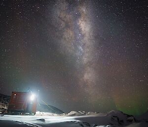 A picture of an orange square field hut at night, with an amazing lit-up milky way and stars overhead