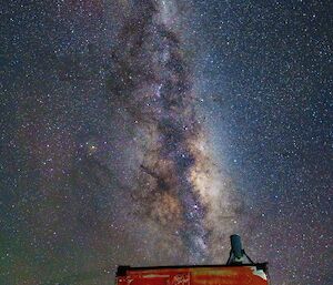 A picture of an orange square field hut at night, with an amazing lit-up milky way and stars overhead