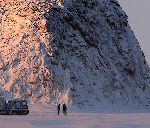 A Hagglands vehicle and two expeditioners on the sea ice, with a large rocky featuring in the immediate background