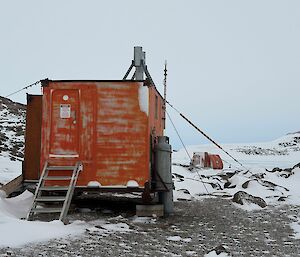 A picture of an orange square field hut, surrounded by snow, rocky outcrops and a cloudy sky