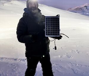 Expeditioner at night with a head-torch on, holding a small solar panel with snow outcrop in background