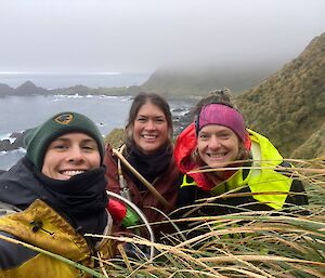 Three women in outdoor gear, standing on a steep tussocky hill with grey hill behind them