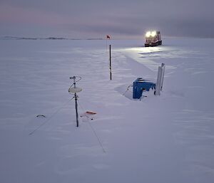 A blue box with an affixed solar panel is being buried by snow, and two poles holding sensors are also visible above the snow