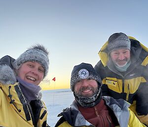 The expeditioners wearing bulky black and yellow jackets and a variety of beanies pose cheerfully with a pale orange sunset visible behind