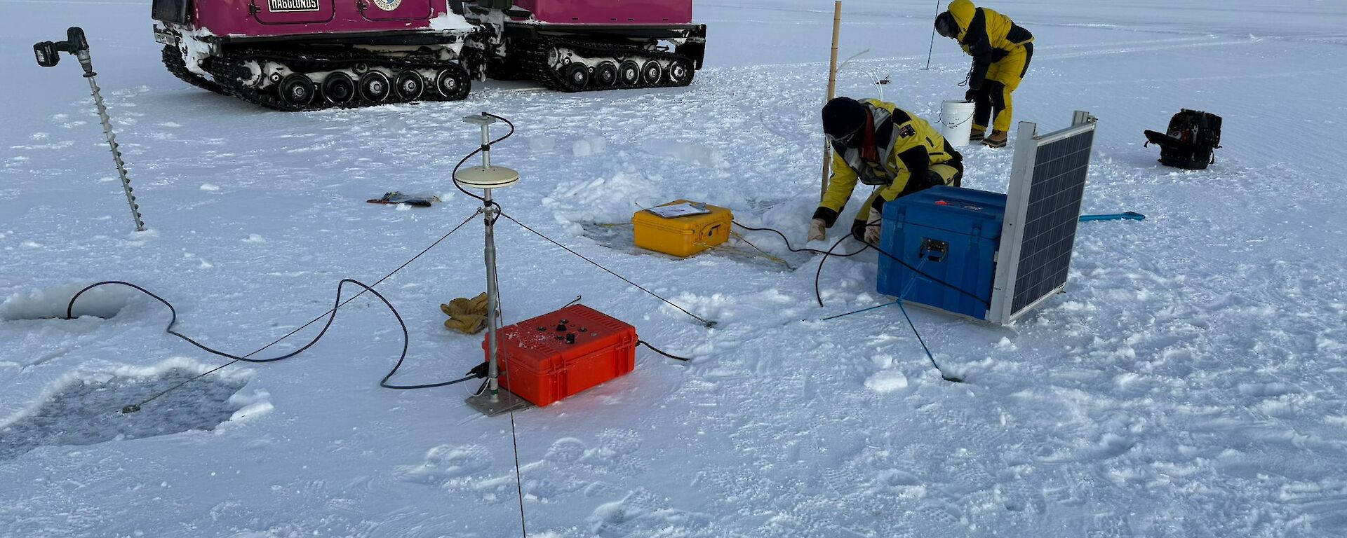 Two people wearing bulky yellow and black jackets work amongst a number of boxes and cables sitting on ice, with a pink Hagglunds vehicle visible behind it and a pale pink dawn sky.