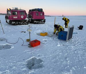 Two people wearing bulky yellow and black jackets work amongst a number of boxes and cables sitting on ice, with a pink Hagglunds vehicle visible behind it and a pale pink dawn sky.