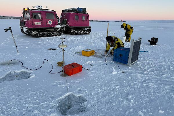 Two people wearing bulky yellow and black jackets work amongst a number of boxes and cables sitting on ice, with a pink Hagglunds vehicle visible behind it and a pale pink dawn sky.