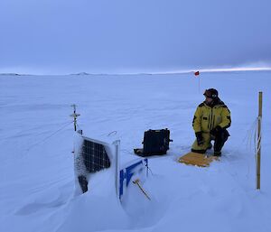 A man wearing a bulky yellow and black jacket kneels next to a laptop in a hard black case, connected to technical equipment sitting on the sea ice.