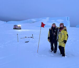 Two people in warm clothing stand proudly next to a red flag and some equipment on the sea ice, with a pink vehicle and a large iceberg visible in the distance