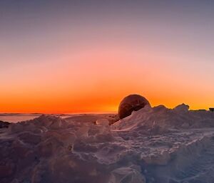 Snowy, uneven surface. The sun rise in the distance and the horizon glows in shades of orange and red against the blue-grey sky.