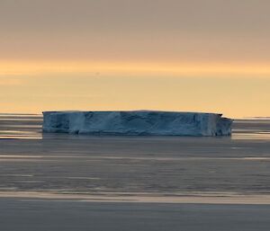 flat, icy ocean with a large iceberg rising out of the water's surface. The horizon is a soft blue with a layer of cloud above.