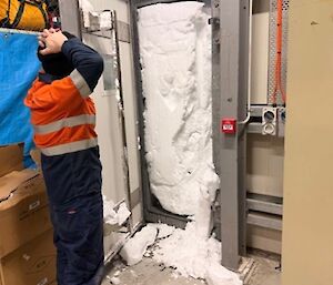 An expeditioner stands with his hands on his head as he looks toward a door with an exit sign above it. The doorway is packed full of snow.