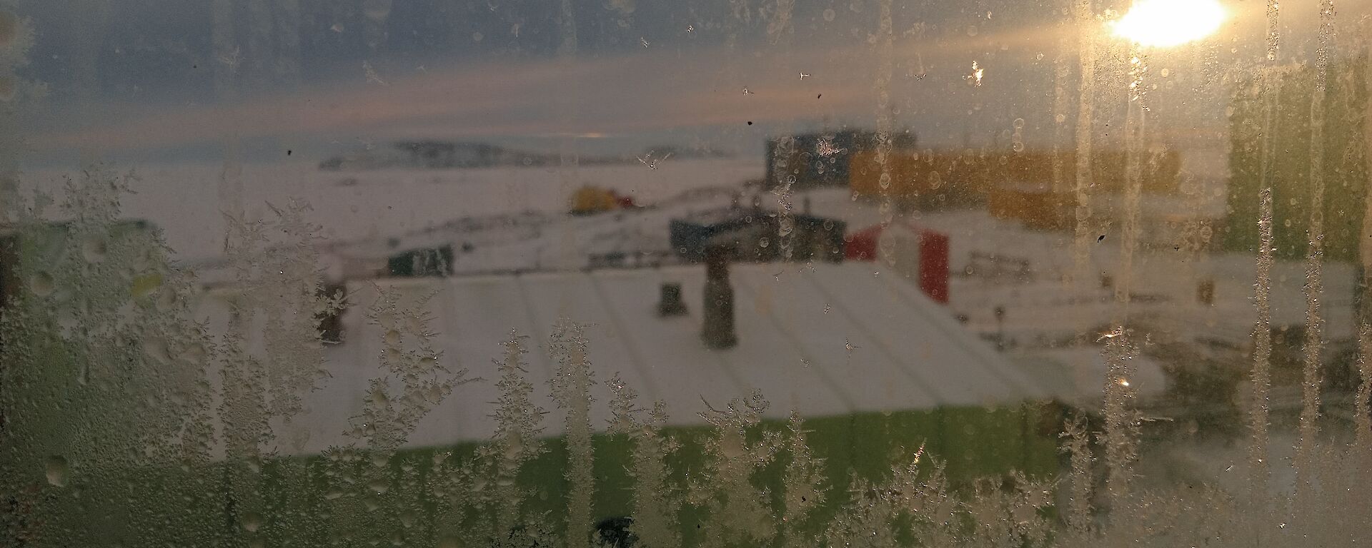 A water front view of the Antarctic Davis station through a bedroom window frosting from the bottom with sun in the background.