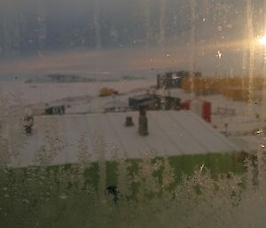A water front view of the Antarctic Davis station through a bedroom window frosting from the bottom with sun in the background.