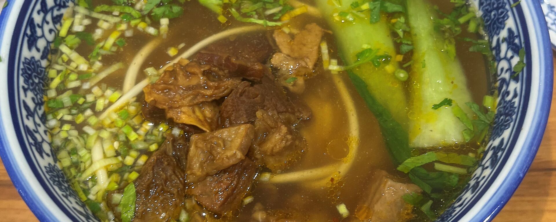 A blue and white bowl filled with broth, beef, vegetables and noodles, rests on a wooden bench.