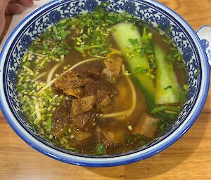 A blue and white bowl filled with broth, beef, vegetables and noodles, rests on a wooden bench.