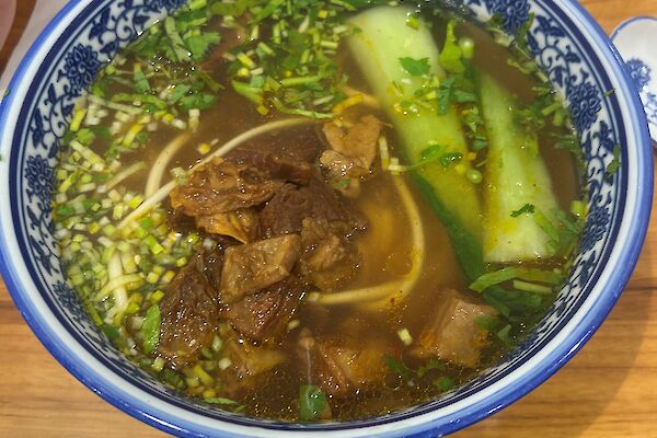 A blue and white bowl filled with broth, beef, vegetables and noodles, rests on a wooden bench.