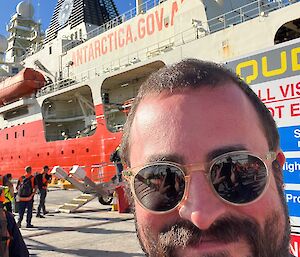 large red and white ship with writing ANTARCTICA.GOV.AU at port behind the smiling face of a bearded man wearing sunglasses.