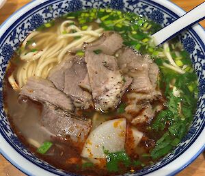 A blue and white bowl filled with broth, beef, vegetables, egg and noodles, rests on a wooden bench.