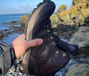 a hand holds a black hiking boot upside down as water pour out of the boot. In the background is the ocean, blue sky and rock shoreline.