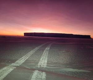 Haggland tracks on the sea ice, moving into the distance where there is a rising sun appearing behind an ice berg