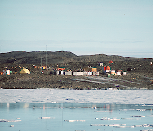A collection of small buildings on a short rise seen over water with pieces of ice floating around