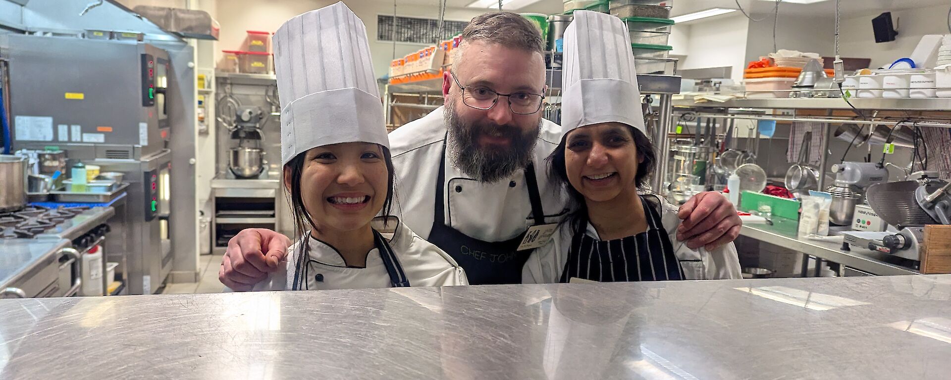 Two women in chef's hats in a kitchen with a smiling chef