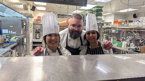 Two women in chef's hats in a kitchen with a smiling chef