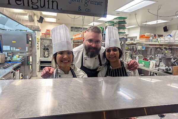 Two women in chef's hats in a kitchen with a smiling chef