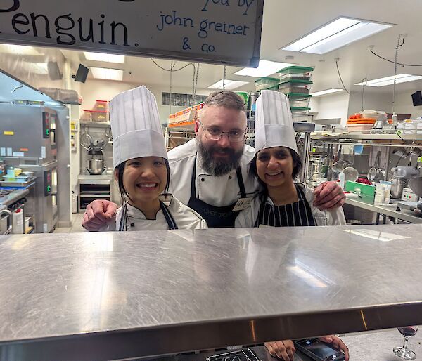 Two women in chef's hats in a kitchen with a smiling chef