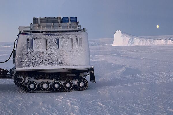 A Hagglands vehicle on the sea ice early morning, overlooking ice cliffs in the background with a bright moon above